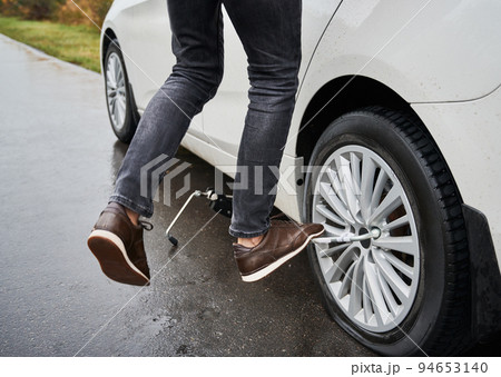 Close up of male auto mechanic standing on metal wrench while unscrewing lug nuts on car wheel. Young man using lug nut wrench while changing flat tire on the road. Concept of emergency road service. Close up of male auto mechanic standing on metal wrench while unscrewing lug nuts on car wheel. Young man using lug nut wrench while changing flat tire on the road. Concept of emergency road service. 94653140