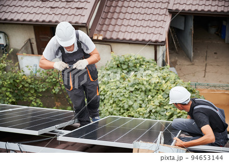 Men workers mounting photovoltaic solar moduls on roof of house. Electrician in helmets wiring solar panels together. Concept of alternative and renewable energy. Men workers mounting photovoltaic solar moduls on roof of house. Electrician in helmets wiring solar panels together. Concept of alternative and renewable energy. 94653144