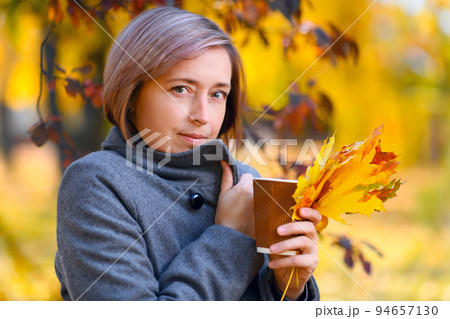 portrait of a beautiful woman with paper cup and colorful yellow leaves, posing in a city park, bright sunny day in autumn 94657130