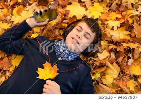 a teenage boy listens to music with headphones, lies on yellow autumn maple leaves, a bright sunny day, beautiful nature in autumn 94657907