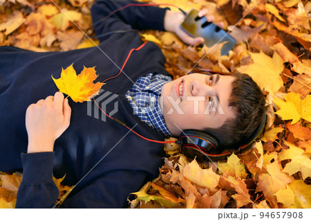 a teenage boy listens to music with headphones, lies on yellow autumn maple leaves, a bright sunny day, beautiful nature in autumn 94657908