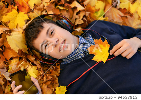 a teenage boy listens to music with headphones, lies on yellow autumn maple leaves, a bright sunny day, beautiful nature in autumn 94657915
