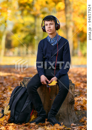 a teenage boy sits in autumn park and listens to music with headphones, trees with yellow leaves on a bright sunny day 94657924