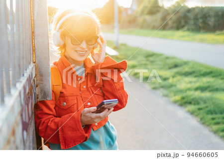 Smiling stylish woman in bright clothes and sunglasses wearing wireless headphones on her head and seting phone for listening music. Walk in city park on sunset. Hipster lifestyle. Selective focus Smiling stylish woman in bright clothes and sunglasses wearing wireless headphones on her head and seting phone for listening music. Walk in city park on sunset. Hipster lifestyle. Selective focus 94660605