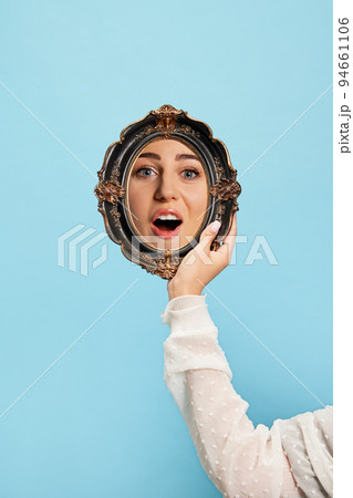 Surprised girl. Woman's hand with retro style mirror with reflection of beautiful girl's face isolated over blue background. Concept of emotions, beauty Surprised girl. Woman's hand with retro style mirror with reflection of beautiful girl's face isolated over blue background. Concept of emotions, beauty 94661106