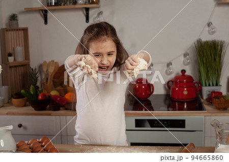 Beautiful little girl dressed in a white blouse is making a dough for baking a bread at a kitchen. Beautiful little girl dressed in a white blouse is making a dough for baking a bread at a kitchen. 94665860