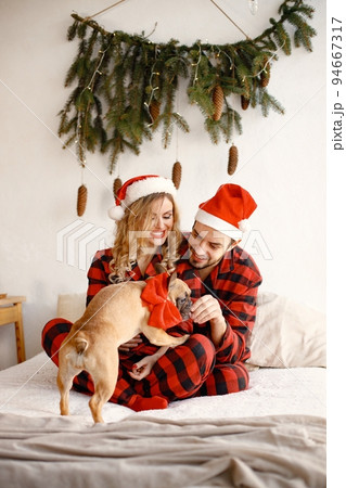 Lovely couple celebrating christmas with dog on a bed. Blonde woman and brunette man wearing plaid pajamas. French bulldog with his family. Lovely couple celebrating christmas with dog on a bed. Blonde woman and brunette man wearing plaid pajamas. French bulldog with his family. 94667317