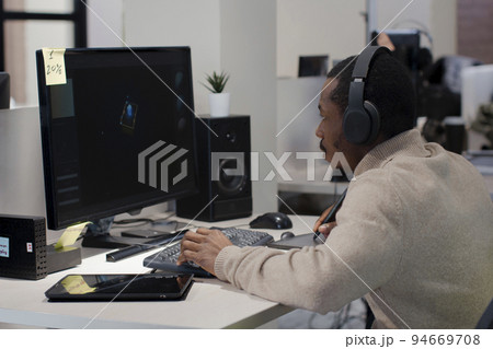 African American designer in headphones draws on tablet 3D models for games. 94669708