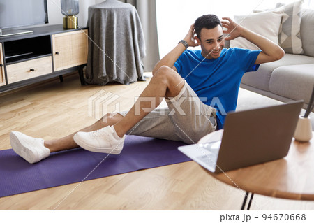 Young arab guy exercising at home, having online fitness video and working on abs strength, looking at laptop screen Young arab guy exercising at home, having online fitness video and working on abs strength, looking at laptop screen 94670668
