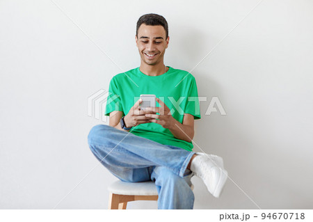 People and technology. Positive arab guy using smartphone sitting on chair over light studio wall background 94670718
