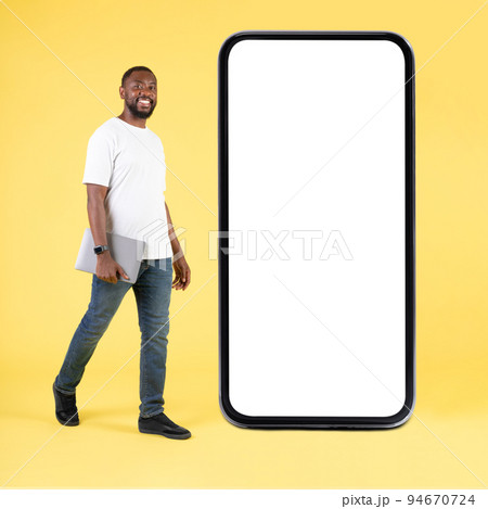 Happy Black Guy Near Large Smartphone Holding Laptop, Yellow Background 94670724