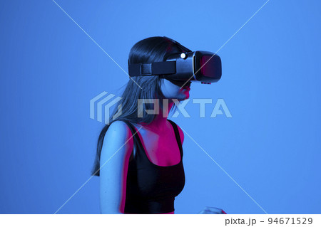 Confident brunette young woman in virtual reality headset pointing in the air while standing in a studio space. Using VR technologies. 94671529