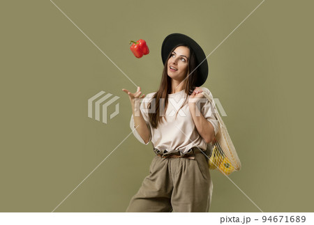 Young excited happy fun vegetarian woman 20s wear casual clothes and black hat hold net eco bag with vegetables after shopping in greengrocer and throw up a red pepper in air. Young excited happy fun vegetarian woman 20s wear casual clothes and black hat hold net eco bag with vegetables after shopping in greengrocer and throw up a red pepper in air. 94671689