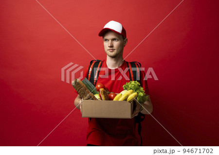 Portrait of smiling cheerful handsome young delivery man holding grocery shopping bag isolated on red background. Portrait of smiling cheerful handsome young delivery man holding grocery shopping bag isolated on red background. 94671702