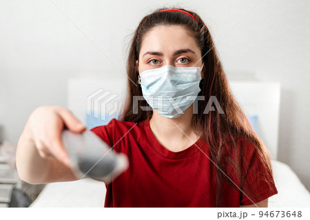 Portrait of a Young woman in a medical mask holding a TV remote control. Close up. The concept of disinformation and false news Portrait of a Young woman in a medical mask holding a TV remote control. Close up. The concept of disinformation and false news 94673648