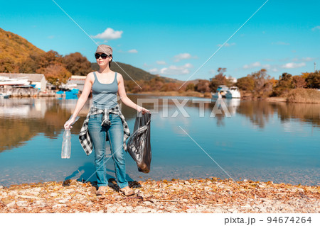 Ecology and garbage collection. Woman volunteer with a garbage bag in his hands, engaged in garbage collection on the lake or sea Ecology and garbage collection. Woman volunteer with a garbage bag in his hands, engaged in garbage collection on the lake or sea 94674264