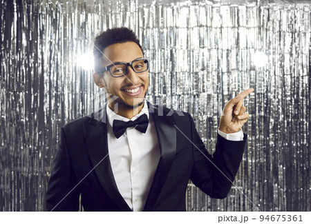 Smiling elegant young man in black tuxedo pointing sideways with index finger on silver background. 94675361