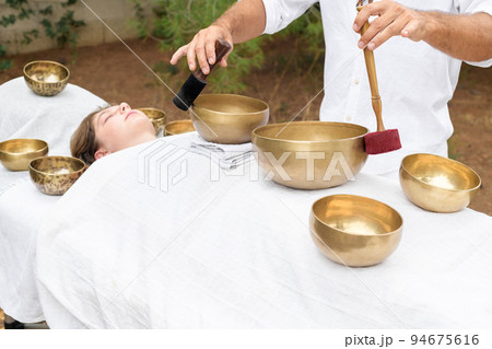Young woman laying on a massage bed with tibetan singing bowl outdoor, male hand holding a felt clapper 94675616