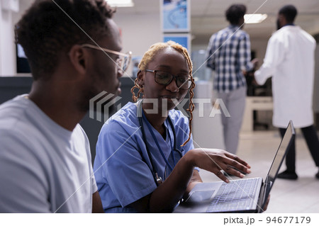 Female nurse using laptop with patient at consultation in waiting area, showing diagnosis results on computer and giving healthcare treatment. Talking to man about disease and medical recovery. Female nurse using laptop with patient at consultation in waiting area, showing diagnosis results on computer and giving healthcare treatment. Talking to man about disease and medical recovery. 94677179