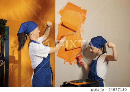 Family using orange color to paint apartment walls at home, painting room together with roller brush and renovating tools. Woman with small child redecorating house, decor improvement. Family using orange color to paint apartment walls at home, painting room together with roller brush and renovating tools. Woman with small child redecorating house, decor improvement. 94677180