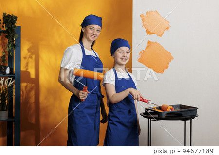 Portrait of family using orange paint color on apartment walls, doing diy home redecoration together. Young kid and mother painting room interior with roller tools and paintbrush. Portrait of family using orange paint color on apartment walls, doing diy home redecoration together. Young kid and mother painting room interior with roller tools and paintbrush. 94677189