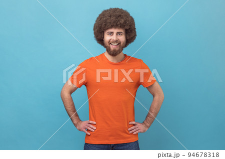 Portrait of cheerful man wearing orange T-shirt looking at camera with toothy smile and happy facial expression, being in good mood, keeps hands on hips. Indoor studio shot isolated on blue background Portrait of cheerful man wearing orange T-shirt looking at camera with toothy smile and happy facial expression, being in good mood, keeps hands on hips. Indoor studio shot isolated on blue background 94678318