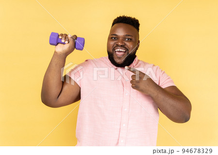 Portrait of excited positive bearded man wearing pink shirt standing with dumbbell and pointing at sport equipment, looking at camera. Indoor studio shot isolated on yellow background. Portrait of excited positive bearded man wearing pink shirt standing with dumbbell and pointing at sport equipment, looking at camera. Indoor studio shot isolated on yellow background. 94678329