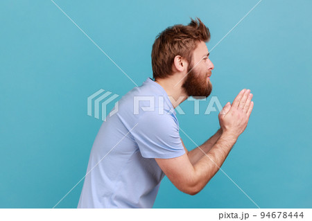 Please, I'm begging. Side view of bearded man holding arms in prayer, asking help or forgiveness with imploring eyes, sincere asking permission. Indoor studio shot isolated on blue background. 94678444