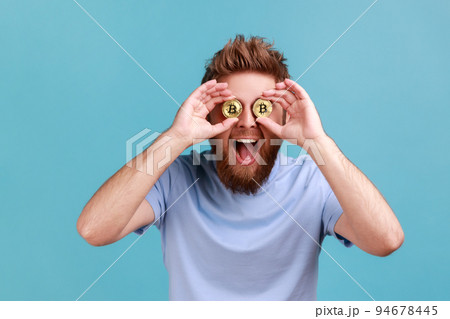 Bitcoin, digital money. Portrait of playful excited bearded man covering eyes with golden bitcoins, being confident in cryptocurrency. Indoor studio shot isolated on blue background. Bitcoin, digital money. Portrait of playful excited bearded man covering eyes with golden bitcoins, being confident in cryptocurrency. Indoor studio shot isolated on blue background. 94678445