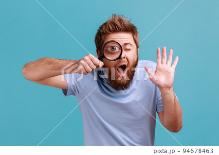 Portrait of funny bearded man holding magnifying glass and looking at camera with big zoom eye, curious face, waving hand, saying hi. Indoor studio shot isolated on blue background. 94678463