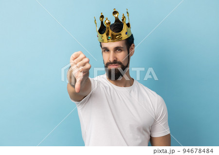 Portrait of sad serious man with beard in white T-shirt and in gold crown standing looking at camera with disappointed expression, showing thumb down. Indoor studio shot isolated on blue background. Portrait of sad serious man with beard in white T-shirt and in gold crown standing looking at camera with disappointed expression, showing thumb down. Indoor studio shot isolated on blue background. 94678484