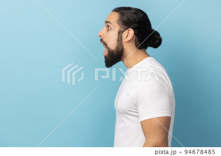 Side view of surprised amazed man with beard wearing white T-shirt looking with shocked expression away, sees something astonishing. Indoor studio shot isolated on blue background. 94678485