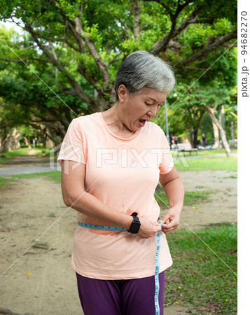 Portrait of happy senior adult elderly asia woman measuring waist with measuring tape in the park. 94682270