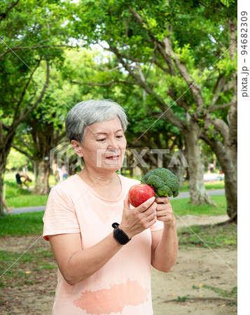 Portrait of happy senior adult elderly asia woman smiling standing and holding fresh vegetable and apple in the park. 94682309