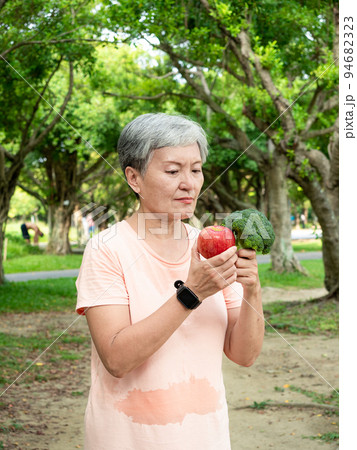Portrait of happy senior adult elderly asia woman smiling standing and holding fresh vegetable and apple in the park. 94682323
