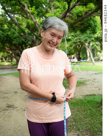Portrait of happy senior adult elderly asia woman measuring waist with measuring tape in the park. 94682344