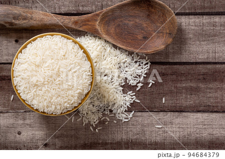 Uncooked dry Thai jasmine rice are overflow from a bowl and wooden ladle beside put on wood floor, angle of view flat lay and top down horizontal view. 94684379