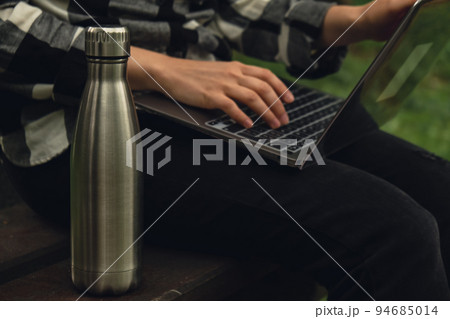 Water bottle. Reusable steel thermo water bottle on wooden bench. Sustainable lifestyle. Plastic free zero waste free living. Go green Woman student work study with laptop in park Water bottle. Reusable steel thermo water bottle on wooden bench. Sustainable lifestyle. Plastic free zero waste free living. Go green Woman student work study with laptop in park 94685014