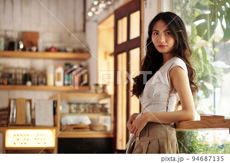 Portrait of serious young woman with long hair standing in coffeeshop and looking at camera 94687135
