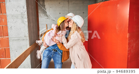 Woman kissing daughter while standing at construction site. Happy parents with child posing in apartment building under construction. Family life and new home purchase concept. Woman kissing daughter while standing at construction site. Happy parents with child posing in apartment building under construction. Family life and new home purchase concept. 94689395