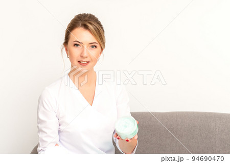 Close-up portrait of young smiling female caucasian healthcare worker standing with jar of cream beauty product on white background. 94690470