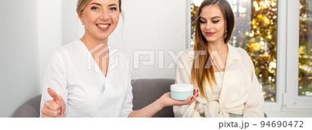 Beautician offering product for young woman, shows thumb up, holding a white plastic jar with a cream, mockup, copy space 94690472
