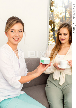 Beautician offering product for the young woman holding a white plastic jar with a cream sitting on the sofa 94690474