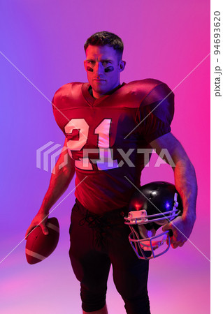 Caucasian male american football player holding helmet and ball with neon pink and blue lighting 94693620