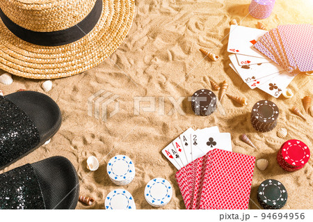 Beachpoker. Chips and cards on the sand. Around the seashells, hat and flip flops. Top view Beachpoker. Chips and cards on the sand. Around the seashells, hat and flip flops. Top view 94694956