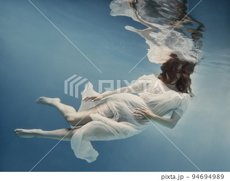 A girl with dark hair in a white dress swims underwater as if in weightlessness 94694989