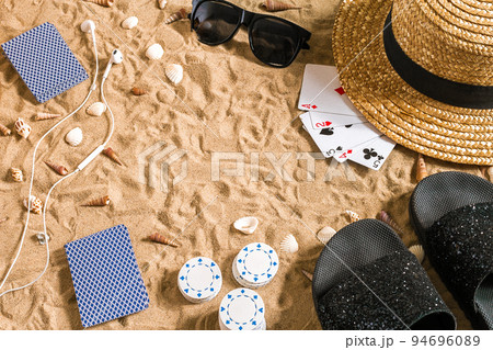 Beachpoker. Chips and cards on the sand. Around the seashells, sunglasses and flip flops. Top view Beachpoker. Chips and cards on the sand. Around the seashells, sunglasses and flip flops. Top view 94696089