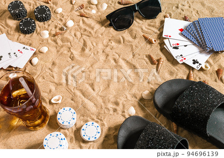 Beachpoker. Chips and cards on the sand. Around the seashells, sunglasses and flip flops. Top view Beachpoker. Chips and cards on the sand. Around the seashells, sunglasses and flip flops. Top view 94696139