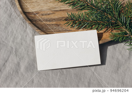 Christmas stationery. Closeup of blank business card, invitation mockup. Green spruce tree branch on wooden tray in sunlight. Grey linen tablecloth. Festive winter still life template. Flat lay, top. Christmas stationery. Closeup of blank business card, invitation mockup. Green spruce tree branch on wooden tray in sunlight. Grey linen tablecloth. Festive winter still life template. Flat lay, top. 94696204