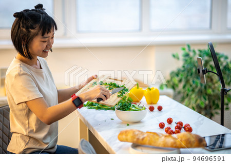 Smiling young housewife cooking in front of the smartphone camera Smiling young housewife cooking in front of the smartphone camera 94696591